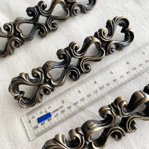 May include: Ornate bronze-toned cabinet pulls with scrolling details and heart-shaped cutouts. The pulls are arranged on a neutral surface with a ruler showing measurements in inches. The pulls have a vintage, antique aesthetic.
