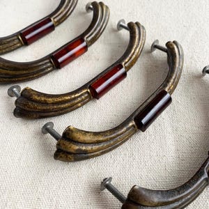May include: Close-up of several antique bronze drawer pulls with a curved design. Each pull features a rectangular, dark red accent in the centre and includes a screw for installation. The pulls have a vintage, aged appearance.