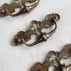 May include: Close-up of several antique brass drawer pulls. Each pull features an ornate, decorative backplate with a curved handle. The pulls have a warm, aged patina, suggesting vintage or retro design.