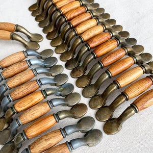 May include: A selection of cabinet pulls with a vintage aesthetic. They combine light brown wooden handles with antique brass-toned metal components. The design incorporates a spoon-shaped end. The hardware is arranged in rows, indicating a large quantity.