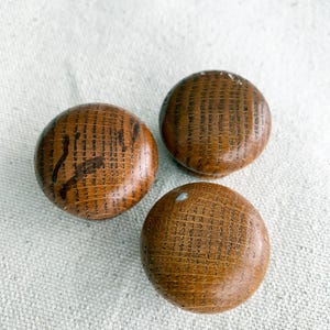 May include: Three round wooden knobs with a warm brown finish and dark grain patterns. The knobs are arranged on a neutral-coloured fabric surface, showcasing their natural wood texture and design. These knobs are suitable for furniture or decorative projects.