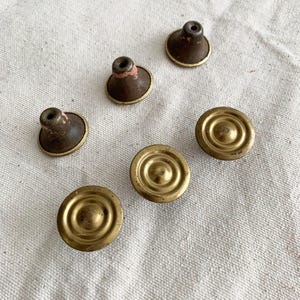 May include: A collection of vintage cabinet knobs. The knobs feature a mix of styles, including round brass knobs with concentric circles and brown knobs with a brass rim. The knobs are arranged on a neutral linen surface.