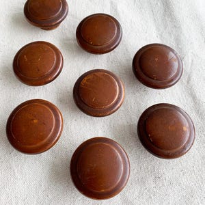 May include: Eight antique brown wooden knobs are displayed. Each knob features a circular design with a raised centre and a rounded edge. The knobs are arranged on a light-coloured surface.