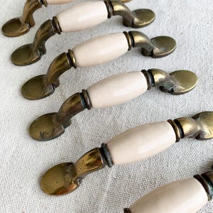 May include: Several antique cabinet pulls with a cream-colored ceramic center and aged brass hardware. The pulls have a curved design and are arranged on a neutral-toned fabric.