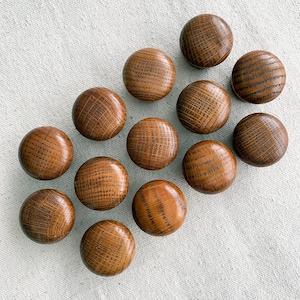 Puede incluir: Una colección de catorce pomos de armario redondos de madera. Los pomos son de color marrón cálido con patrones de vetas de madera visibles. Los pomos están dispuestos en diagonal sobre una superficie de tela de color claro.