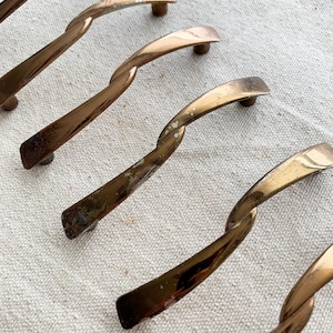 Może przedstawiać: Zbliżenie na kilka uchwytów do szafek z mosiądzu w stylu vintage. Każdy uchwyt ma skręconą konstrukcję i zakrzywiony uchwyt. Mosiądz ma ciepłą, postarzaną patynę z pewnymi śladami zużycia, co dodaje im uroku.