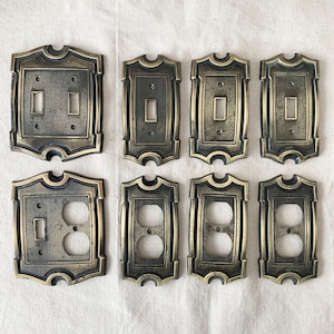 Può includere: Otto placche per interruttori elettrici color ottone antico. Le placche hanno un design decorativo e ornato con un centro rettangolare per interruttori e prese. Le placche sono in varie configurazioni, inclusi stili a interruttore e presa singola e doppia.