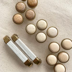 May include: A selection of wooden cabinet knobs and pulls. The knobs feature a round, light-coloured wood centre with a brass-coloured metal rim. The pulls are white with a wood grain design and brass-coloured metal ends. The items are arranged on a neutral-coloured fabric.
