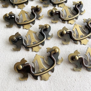Könnte beinhalten: Eine Sammlung antiker Schubladengriffe aus Messing. Jeder Griff hat eine dekorative Rückplatte mit einem verzierten Design und einem gebogenen Griff. Das Messing hat eine warme, gealterte Patina.