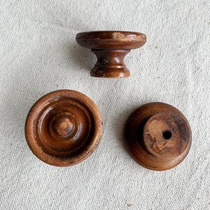 May include: Three antique wooden furniture knobs in varying shapes. One knob is a small pedestal shape, while the other two are circular with a central knob. The knobs are a warm brown color and show signs of wear.