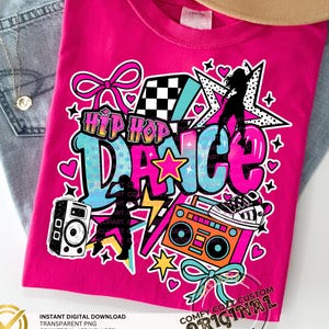 May include: A bright pink t-shirt with a colourful "Hip Hop Dance" graphic. The design includes a boombox, speaker, stars, hearts, and dancer silhouettes. The t-shirt is displayed on a pair of blue jeans.