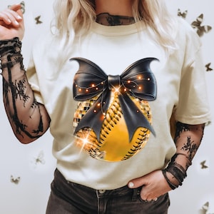 May include: A cream-colored short-sleeve t-shirt featuring a graphic print of a yellow softball partially covered in gold disco ball tiles. A large black bow with thin string lights is positioned over the top of the softball graphic.
