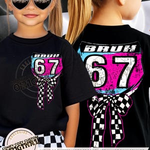 Puede incluir: Camiseta negra con un diseño de placa de motocross rosa, azul y blanco con el número 67 y la palabra "BRUH". El diseño incluye un lazo a cuadros. La camiseta forma parte de una colección "Comfy Cozy Custom Original Design".