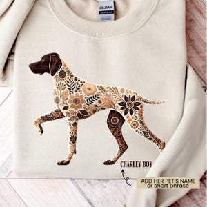 May include: Cream-colored sweatshirt featuring a floral-patterned dog design in shades of brown and tan. The text "CHARLEY BOY" is below the dog, with an arrow pointing to it. The sweatshirt is a Gildan Heavy Blend.
