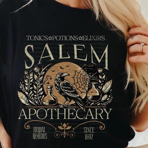 May include: Black t-shirt featuring a Salem Apothecary design. The graphic includes a raven, potion bottles, a spiderweb, and the text "TONICS POTIONS ELIXIRS" and "HERBAL REMEDIES SINCE 1692". The design has a vintage, gothic aesthetic.
