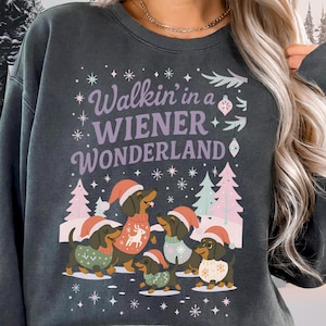Puede incluir: Sudadera gris oscuro con el texto "Walkin' in a WIENER Wonderland". Presenta un gráfico de perros salchicha con gorros y suéteres de Papá Noel, rodeados de copos de nieve y árboles de Navidad. Un diseño festivo navideño.