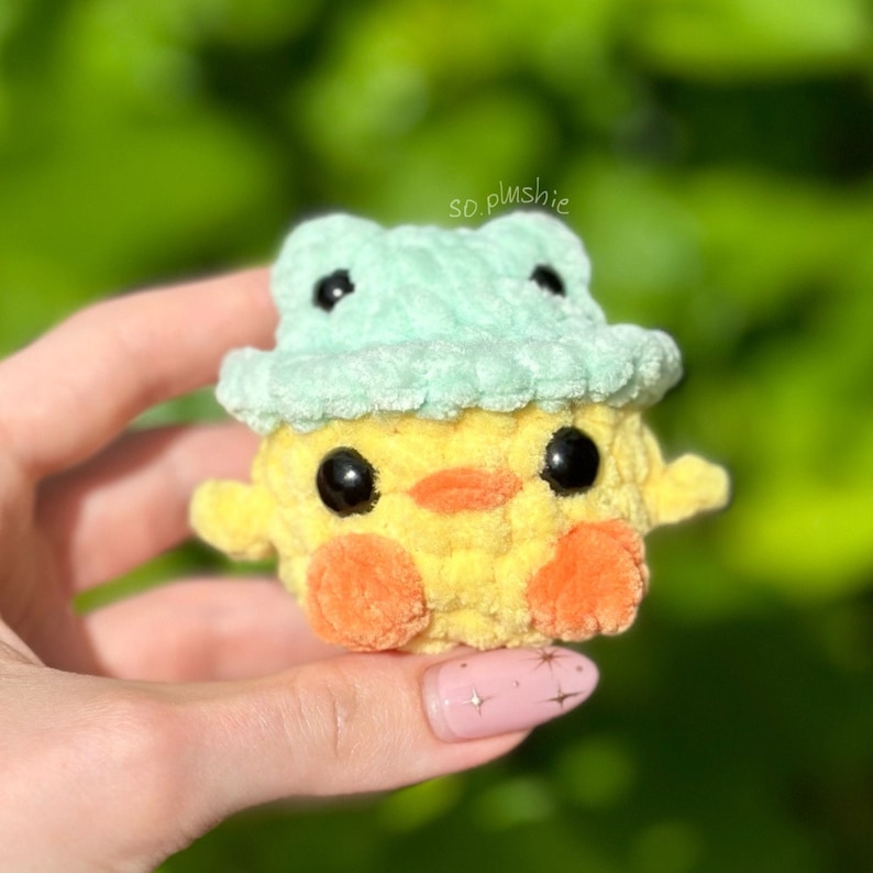 May include: A yellow crocheted chick wearing a light green crocheted frog hat. The chick has black eyes and orange feet.