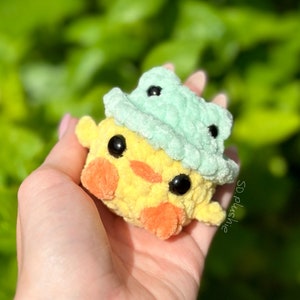 May include: A small yellow crocheted chick wearing a light green crocheted hat. The chick has black eyes and an orange beak. The chick is being held in a hand.
