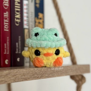 May include: A yellow crocheted chick wearing a green crocheted frog hat. The chick has black eyes and an orange beak. The chick is sitting on a wooden shelf.