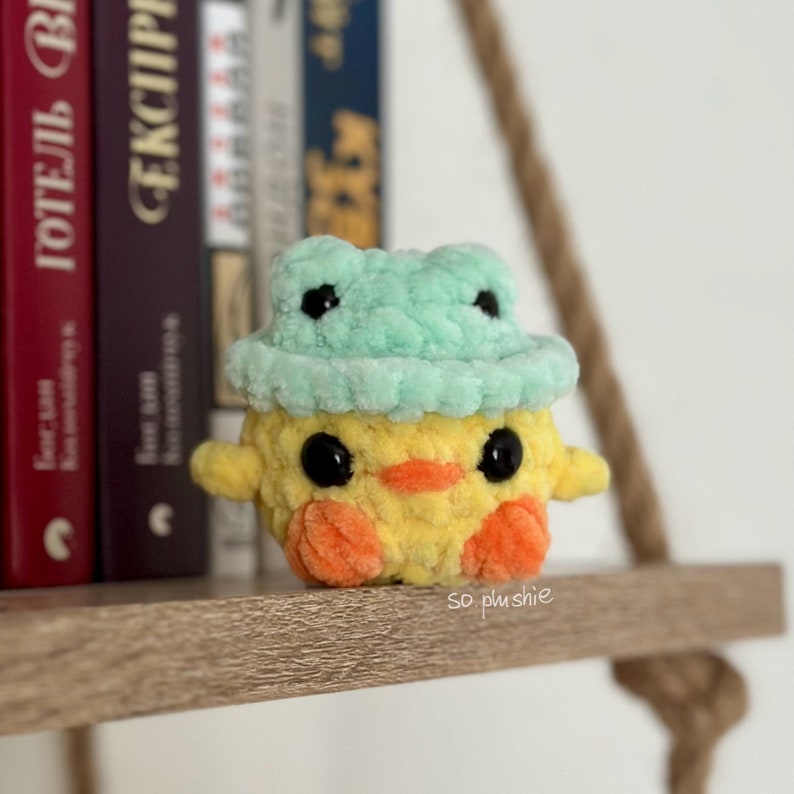 May include: A yellow crocheted chick wearing a green crocheted frog hat. The chick has black eyes and an orange beak. The chick is sitting on a wooden shelf.