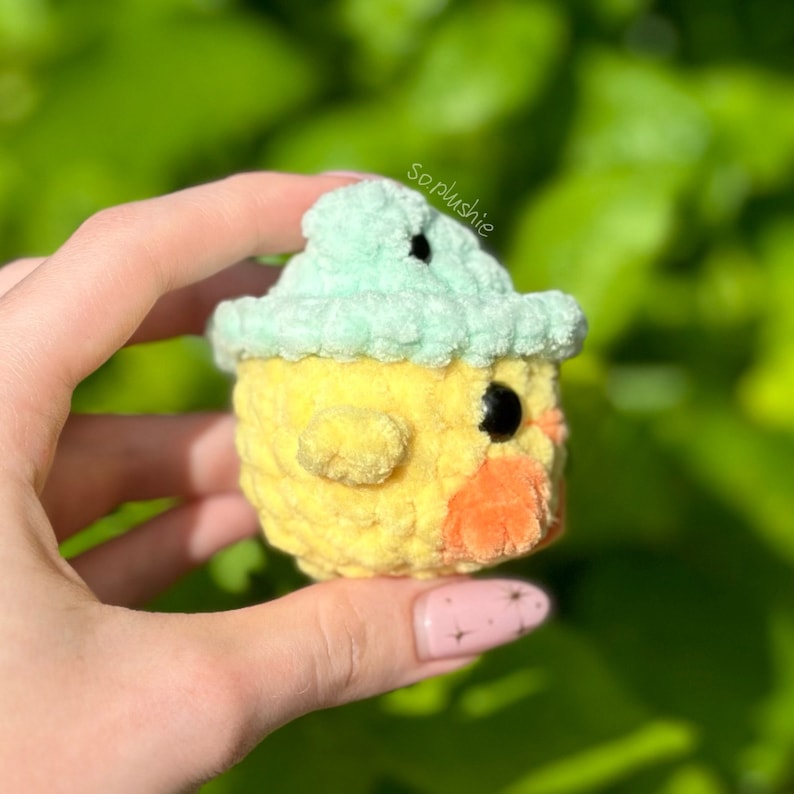 May include: A yellow crocheted chick wearing a light green crocheted hat. The chick has a small orange beak and black eyes.