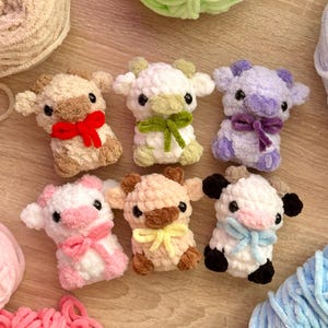 Può includere: Sei peluche di mucca all'uncinetto fatti a mano in vari colori, ognuno con un fiocco. Le mucche sono nei toni del marrone, bianco e viola, con fiocchi rossi, verdi, rosa, gialli e blu. Sullo sfondo ci sono gomitoli di lana nei colori coordinati.