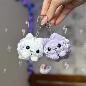 May include: Two fluffy crocheted cat keychains, one white and one purple, with black eyes and a pink nose. The keychains are attached to a silver key ring.