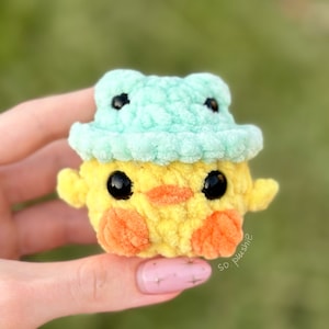 May include: A small crocheted yellow chick wearing a light blue crocheted frog hat. The chick has black eyes and an orange beak. The hat has two green frog eyes.