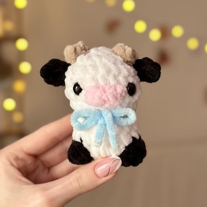 May include: A small crocheted cow plush toy with white and black fur, pink nose, and black eyes. The cow is wearing a blue bow.