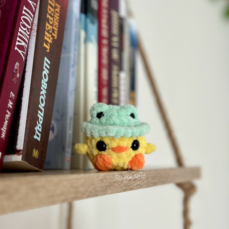 May include: A yellow crocheted duck wearing a green crocheted frog hat. The duck has black eyes, an orange beak, and orange feet. The duck is sitting on a wooden shelf.