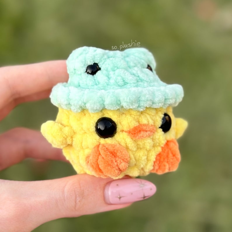 May include: A small yellow crocheted chick wearing a light blue crocheted hat. The chick has black eyes and an orange beak.
