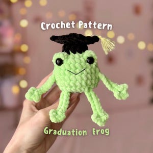May include: A light green crochet frog wearing a black graduation cap with a yellow tassel. The frog has large black eyes and a stitched smile. Text on the image reads "Crochet Pattern" and "Graduation Frog".