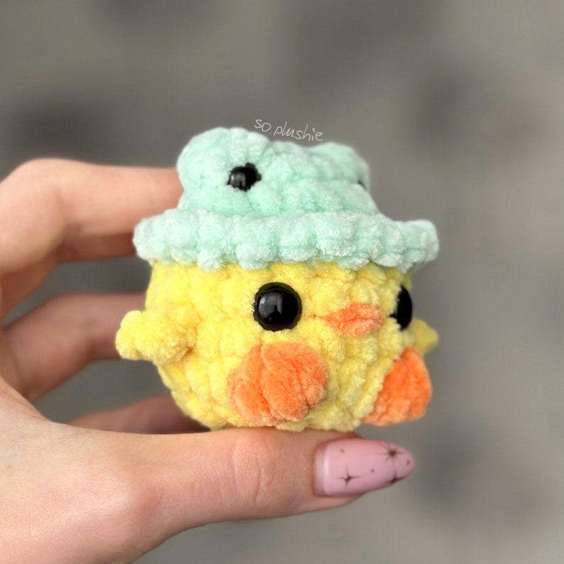 May include: A small yellow crocheted chick wearing a light green crocheted hat with black eyes. The chick is holding a small orange carrot in its beak.
