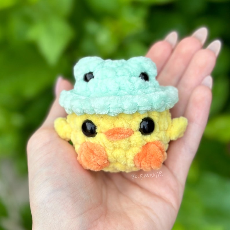 May include: A yellow crocheted chick wearing a light blue crocheted frog hat. The chick has black eyes and an orange beak. The chick is sitting in the palm of a hand.