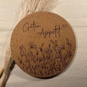 Könnte beinhalten: Ein runder, brauner Korkuntersetzer mit dem Schriftzug "Guten Appetit", einem Herz und einem floralen Design. Der Untersetzer liegt auf einem cremefarbenen Stoff, im Hintergrund getrocknetes Ziergras.