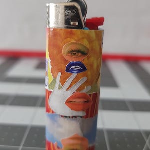 May include: A red, white, and blue lighter with a collage of eyes, lips, and a hand on the side. The lighter is made of metal and has a silver top.