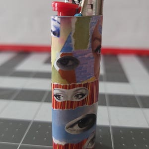 May include: A red, white, and blue lighter with a collage of eyes on the body. The lighter has a silver top and a red button.