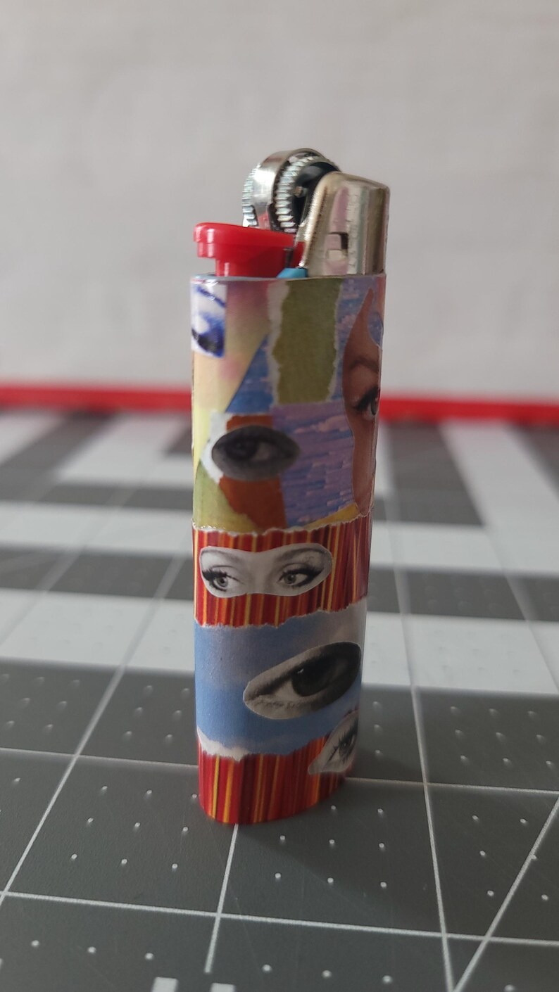 May include: A red, white, and blue lighter with a collage of eyes on the body. The lighter has a silver top and a red button.