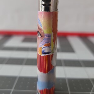 May include: A silver lighter with a colorful collage design featuring a woman's eye, abstract shapes, and stripes. The design is reminiscent of a vintage magazine cover.