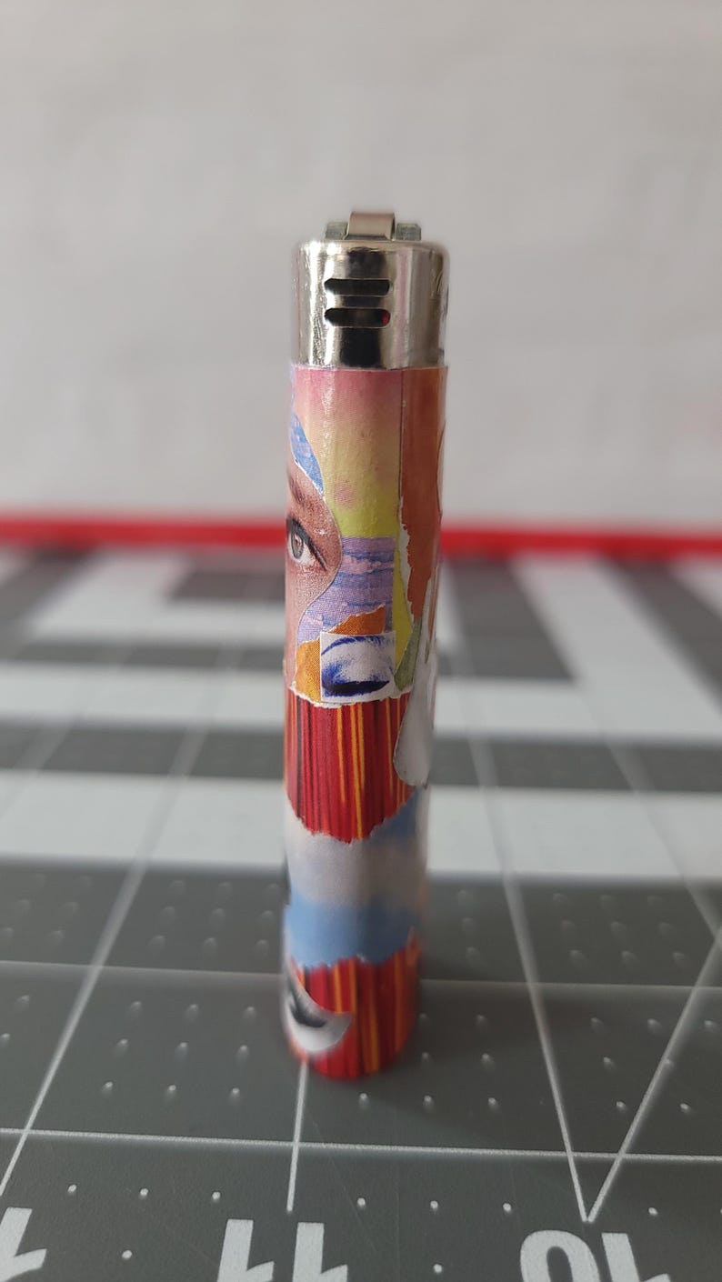 May include: A silver lighter with a colorful collage design featuring a woman's eye, abstract shapes, and stripes. The design is reminiscent of a vintage magazine cover.
