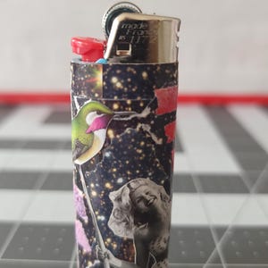 May include: A black and silver lighter with a colorful collage design featuring a hummingbird, a woman, and a galaxy-like background. The lighter has the text "made in France 8 11 72" printed on the side.