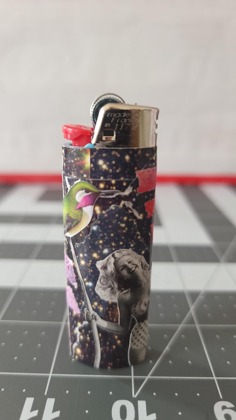 May include: A black and silver lighter with a colorful collage design featuring a hummingbird, a woman, and a galaxy-like background. The lighter has the text "made in France 8 11 72" printed on the side.
