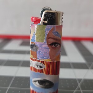 May include: A silver lighter with a red top and a colorful design featuring multiple eyes. The lighter is made in France and has the number 1172 printed on it.