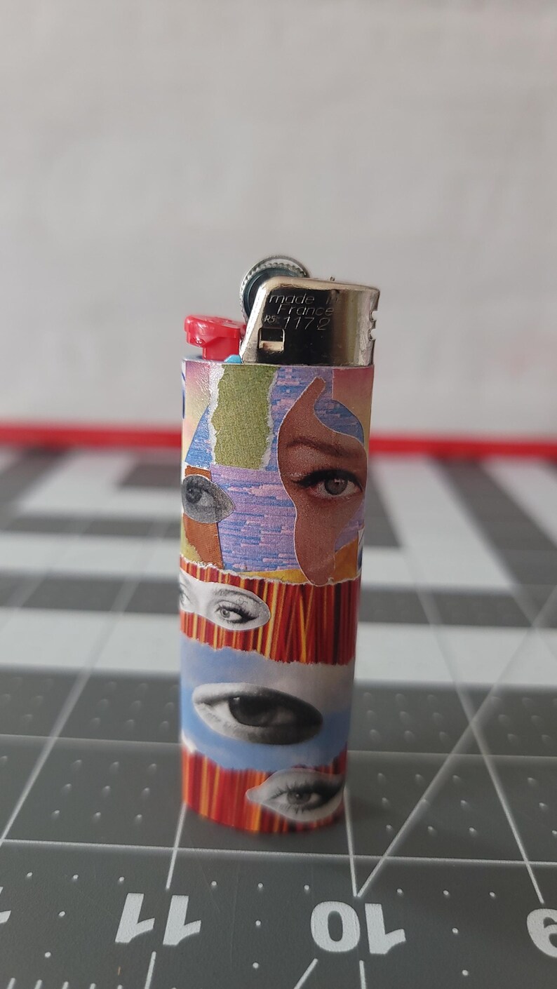 May include: A silver lighter with a red top and a colorful design featuring multiple eyes. The lighter is made in France and has the number 1172 printed on it.