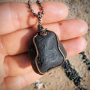 Puede incluir: Un colgante de piedra negra envuelto en alambre de cobre, colgando de una cadena de cobre. El colgante es una piedra natural con una textura rugosa.