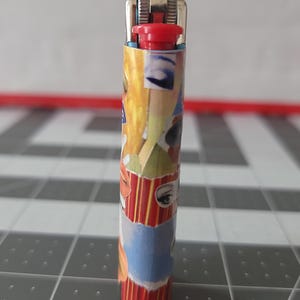 May include: A red, white, and blue lighter with a collage of images on the side. The images include eyes, lips, and other abstract designs.