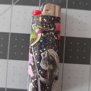 May include: A black lighter with a colorful collage design featuring a hummingbird, a woman, and a starry sky background.