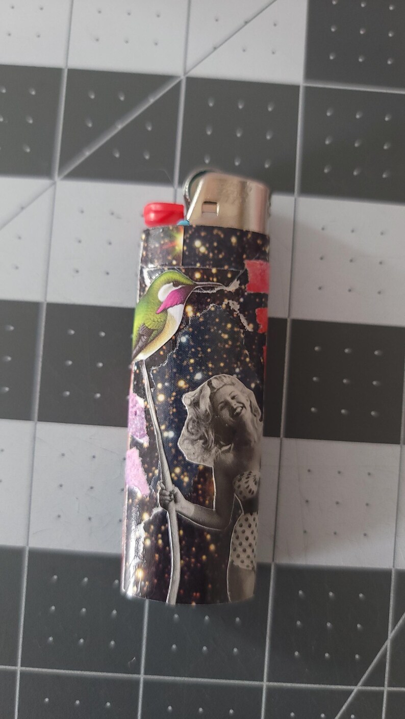 May include: A black lighter with a colorful collage design featuring a hummingbird, a woman, and a starry sky background.