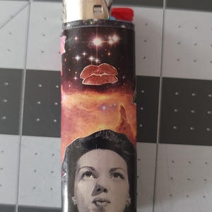 May include: A black lighter with a colorful design featuring a woman's face, a pair of lips, and a galaxy-like background.