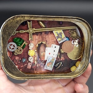 Puede incluir: Una pequeña lata redonda de metal con tapa de vidrio. Dentro de la lata hay varios objetos, incluyendo una espada, cartas de juego, una calavera, una moneda y una carta de tarot. La lata está decorada con un borde dorado.